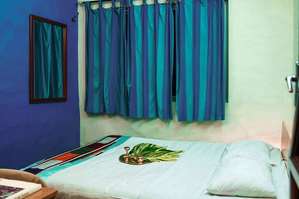 Cozy bedroom in IVAC Ayurvedic Centre India with a simple double bed, adorned with a colourful runner and a decorative tray with leaves. The room features blue-striped curtains and a mirror on the wall, creating a relaxing atmosphere.