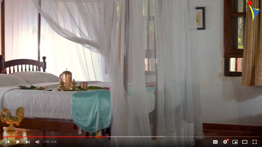 Elegant room at IVAC Ayurvedic Centre India featuring a cozy bed with white linens, adorned with brass Ayurveda accessories, surrounded by sheer white curtains.