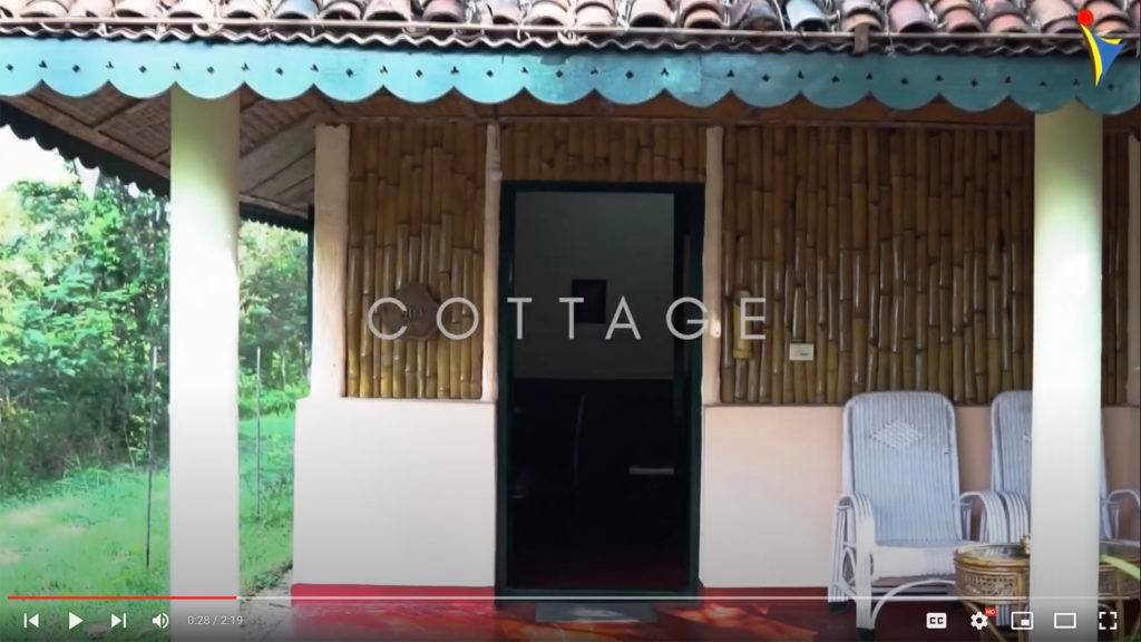Exterior view of a cottage at IVAC Ayurvedic Centre India, featuring bamboo walls, a tiled roof, and a small porch with wicker chairs.