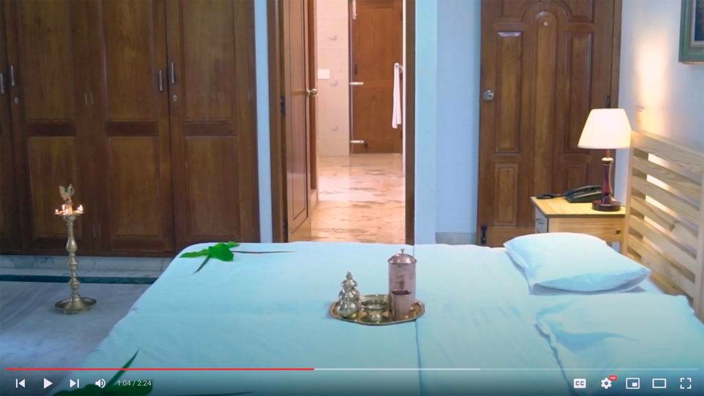 Interior of a room at IVAC Ayurvedic Centre in India, featuring a bed with blue sheets, traditional brass lamp, and decorative items on tray; wooden furniture and warm lighting enhance the serene atmosphere.