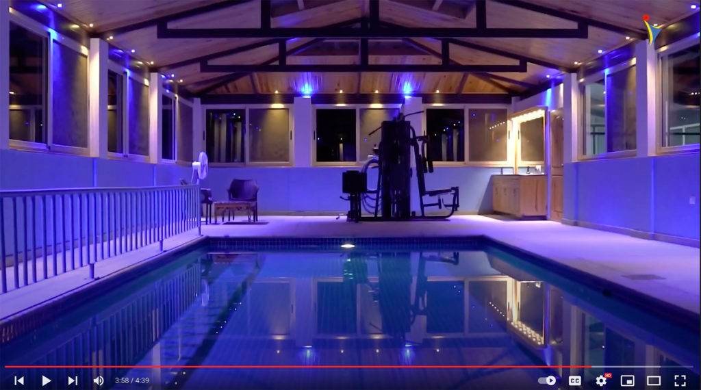 Indoor swimming pool area at IVAC Ayurvedic Centre India featuring wooden ceiling with blue accent lighting, lounge chairs, and gym equipment near windows for relaxation and exercise.