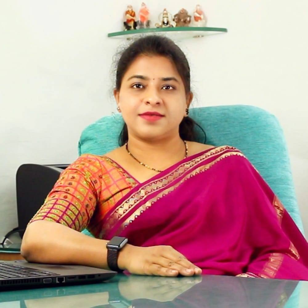 Woman in a traditional Indian sari sitting at a desk, with a laptop and spiritual figurines on a shelf behind her, related to Ayurvedic wellness.