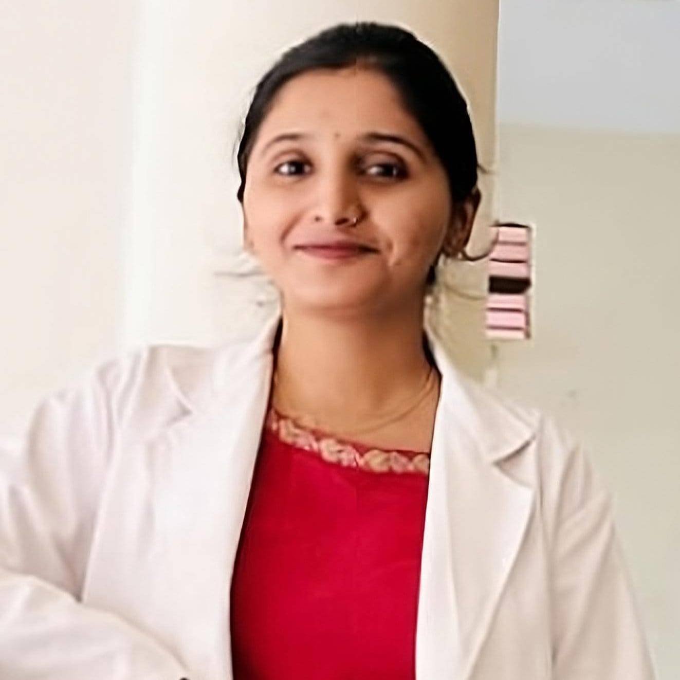 Portrait of a woman in a white coat and red attire, smiling confidently. The setting is a light-colored room, relating to Ayurvedic wellness and Panchakarma.