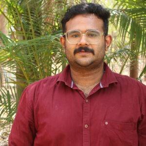 A person wearing glasses and a maroon shirt stands in front of green foliage, related to Ayurvedic wellness and Panchakarma.