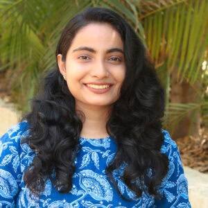 Portrait of a young woman with long, curly black hair wearing a blue floral patterned top, smiling at the camera, with green foliage in the background, representing Ayurvedic wellness.