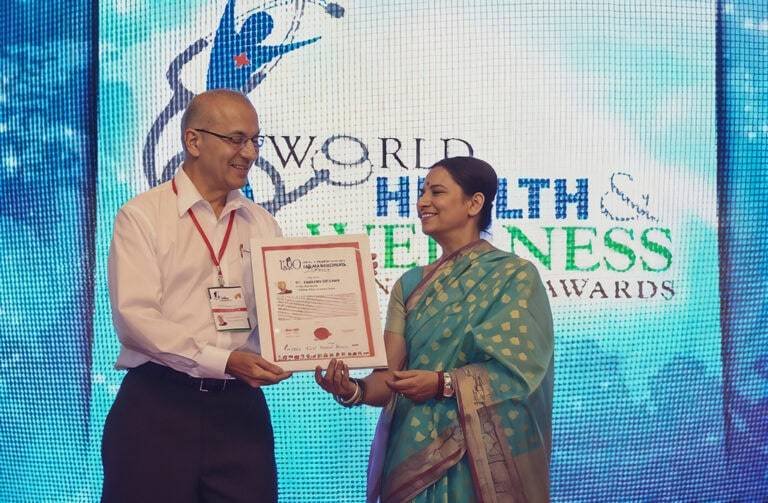 Dr. Talavane Krishna receiving an award at the World Health and Wellness Awards ceremony. Dr. Krishna, dressed in formal attire, is seen smiling and graciously accepting the award certificate from a distinguished presenter in a traditional saree. The backdrop displays the event’s logo, emphasizing the global recognition of excellence in health and wellness. This moment captures Dr. Krishna’s dedication to advancing Ayurvedic practices and his significant contributions to the field of holistic healing.