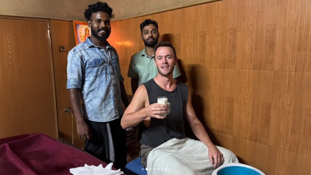 A man wearing a sleeveless shirt sits in a wooden-paneled room, smiling at the camera while holding a glass of milky liquid. Two other men stand behind him, and a small bucket is placed near his seat.